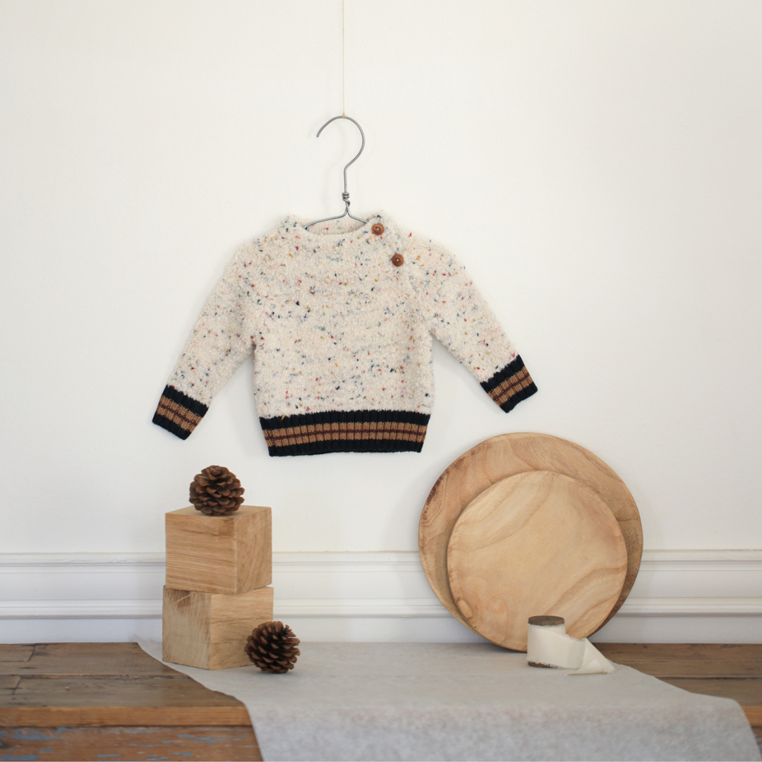 Sold at Bombon shop, designed by 1+ in the family — speckled knit baby sweater with contrast ribbed cuffs and buttoned shoulder, in cream with navy and brown accents. lifestyle