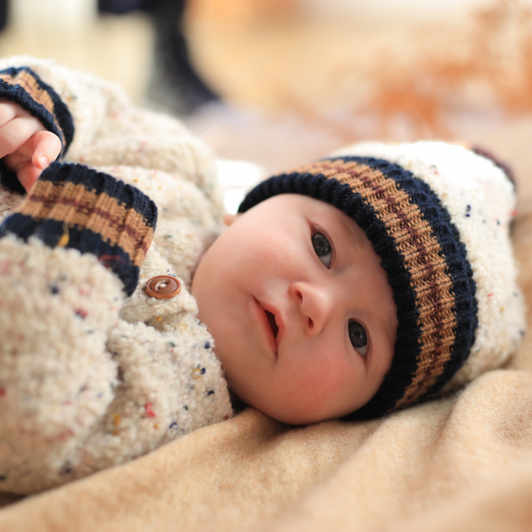 Sold at Bombon shop, designed by 1+ in the family — speckled knit baby sweater with contrast ribbed cuffs and buttoned shoulder, in cream with navy and brown accents. model