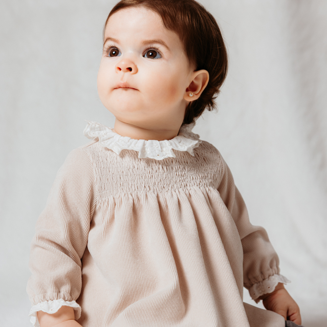 Sold at Bombon shop, designed by Fina Ejerique, a beige corduroy baby dress with smocked bodice, white ruffle collar, and long sleeves. model