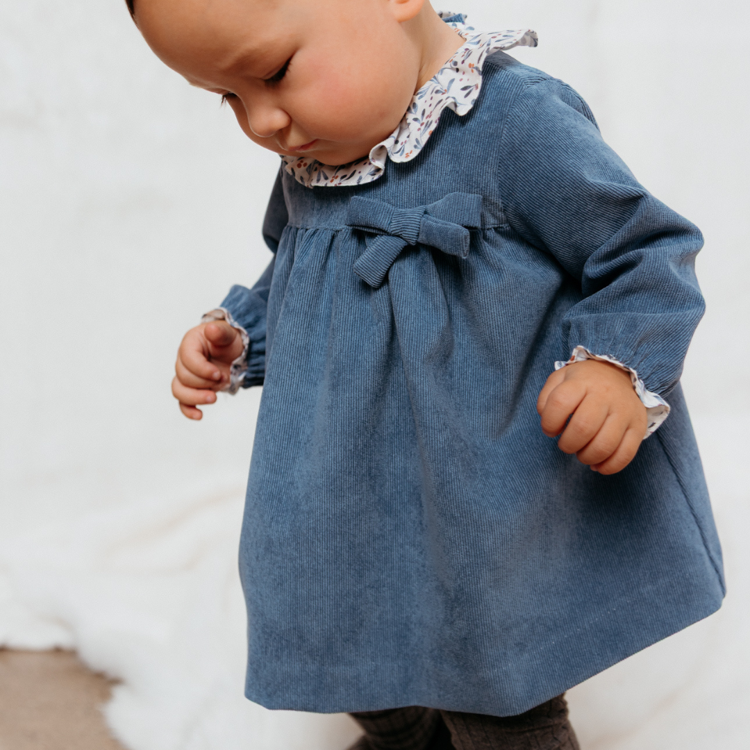 Sold at Bombon shop, designed by Fina Ejerique, a sky-blue corduroy baby dress with floral ruffle collar, bow detail, and long sleeves. model
