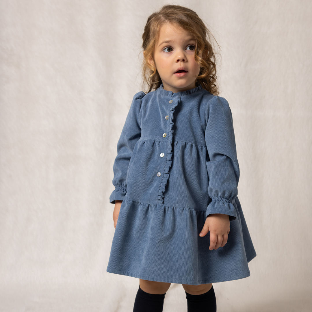 Sold at Bombon shop, designed by Fina Ejerique, a blue corduroy baby dress with ruffle trim, button front, and long sleeves. model