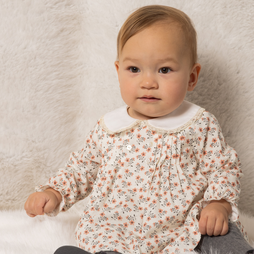 Sold at Bombon shop, designed by Fina Ejerique, a white viyella baby dress with pink and green floral print, Peter Pan collar with lace trim, pleated front, and long sleeves. model