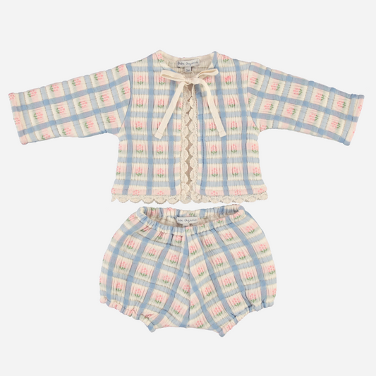 Sold at Bombon Shop, this Bebe Organic baby girl outfit features a charming pastel plaid pattern in soft blue, pink, and cream tones, with a long-sleeve top adorned with lace trim, scalloped edges, and a delicate front tie, paired with matching bloomers — a vintage-inspired set made from organic cotton for comfort and timeless style.
