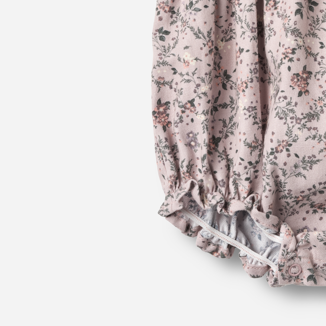 Sold at Bombon shop, designed by Wheat — sleeveless lilac floral baby romper with ruffled shoulders, gathered waist, and elasticized leg openings with snap closures for easy changing. bottom