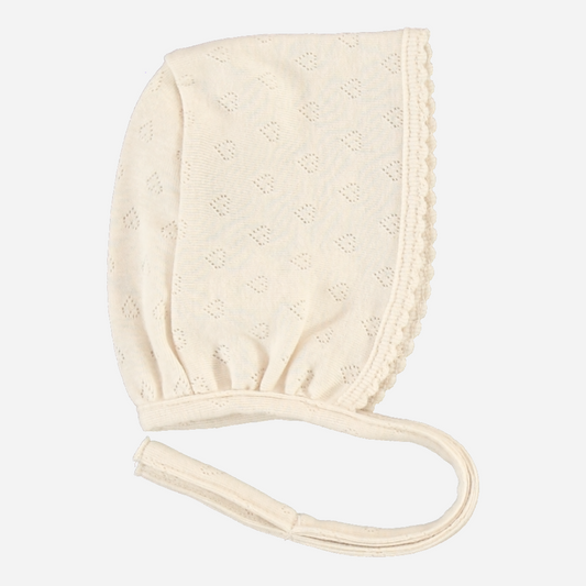 Sold at Bombon Shop, this Bebe Organic baby bonnet is made from soft organic cotton in a delicate ivory pointelle knit, featuring scalloped trim and secure ties — pair with the matching footed romper for a timeless and cozy newborn look perfect for first outings and gifting.