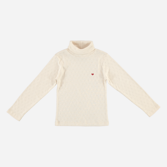 Sold at Bombon Shop, this Bebe Organic girls’ turtleneck top is crafted from soft organic cotton in an ivory pointelle knit, featuring long sleeves, a high neckline for warmth, and a subtle embroidered red heart — a cozy layering essential with a sweet, minimalist touch.