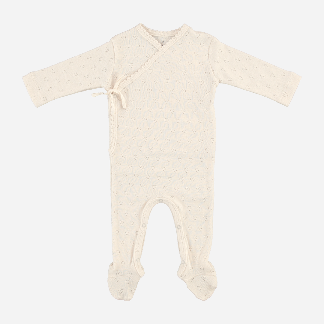Sold at Bombon Shop, this Bebe Organic newborn footed romper is crafted from ultra-soft organic cotton in a delicate ivory pointelle knit, featuring a wrap-style front with side tie and snap closures for easy changing — a gentle and timeless essential for baby’s first wardrobe.