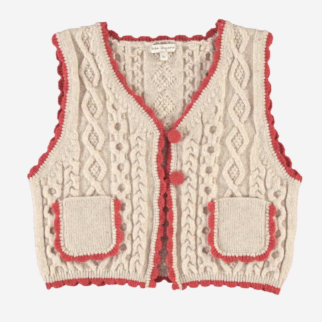 Sold at Bombon Shop, this Bebe Organic cable knit vest for baby and toddler girls is crafted from soft organic cotton in a natural beige tone, featuring red scalloped trim, front patch pockets, and pom-pom button details — a cozy layering piece with vintage charm and sustainable style.