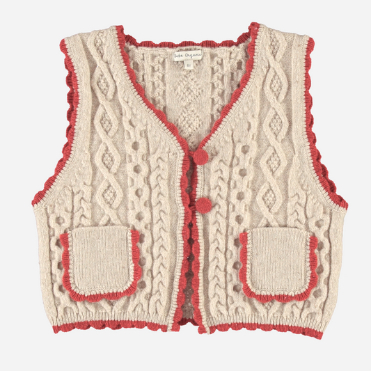 Sold at Bombon Shop, this Bebe Organic cable knit vest for baby and toddler girls is crafted from soft organic cotton in a natural beige tone, featuring red scalloped trim, front patch pockets, and pom-pom button details — a cozy layering piece with vintage charm and sustainable style.