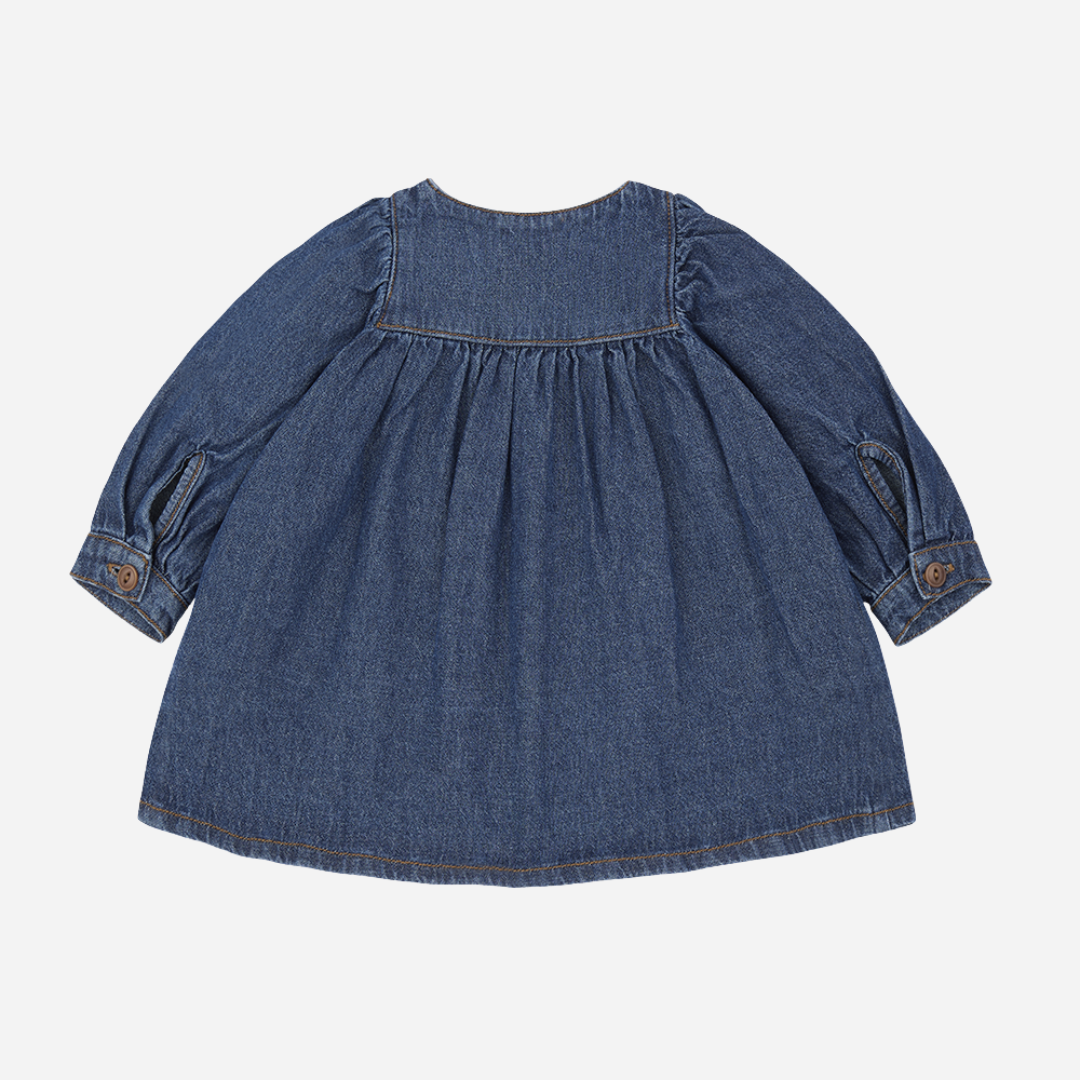 Sold at Bombon shop, designed by 1+ in the family — soft denim baby dress with long sleeves, wooden buttons, and gathered waist. back