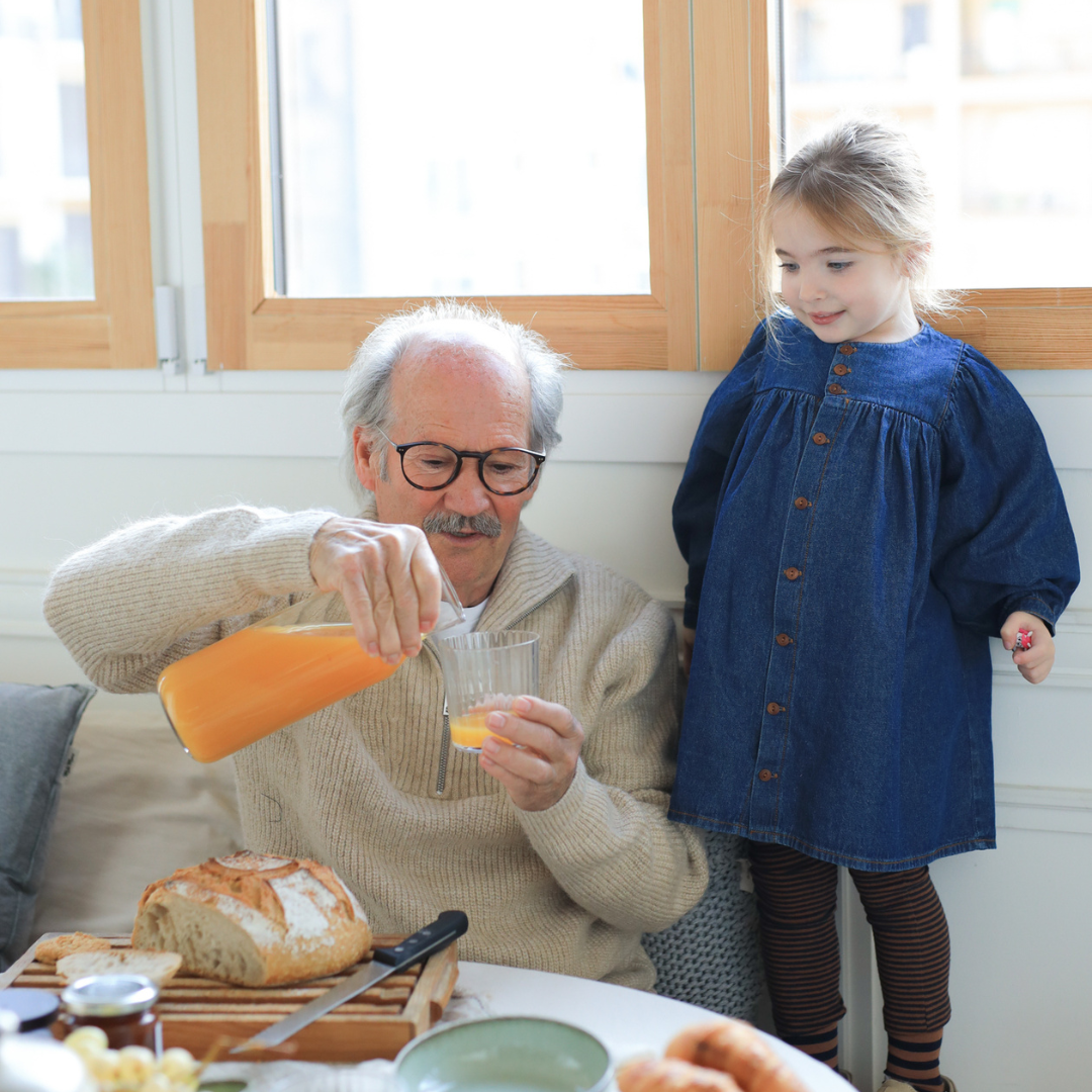 Sold at Bombon shop, designed by 1+ in the family — soft denim baby dress with long sleeves, wooden buttons, and gathered waist. model
