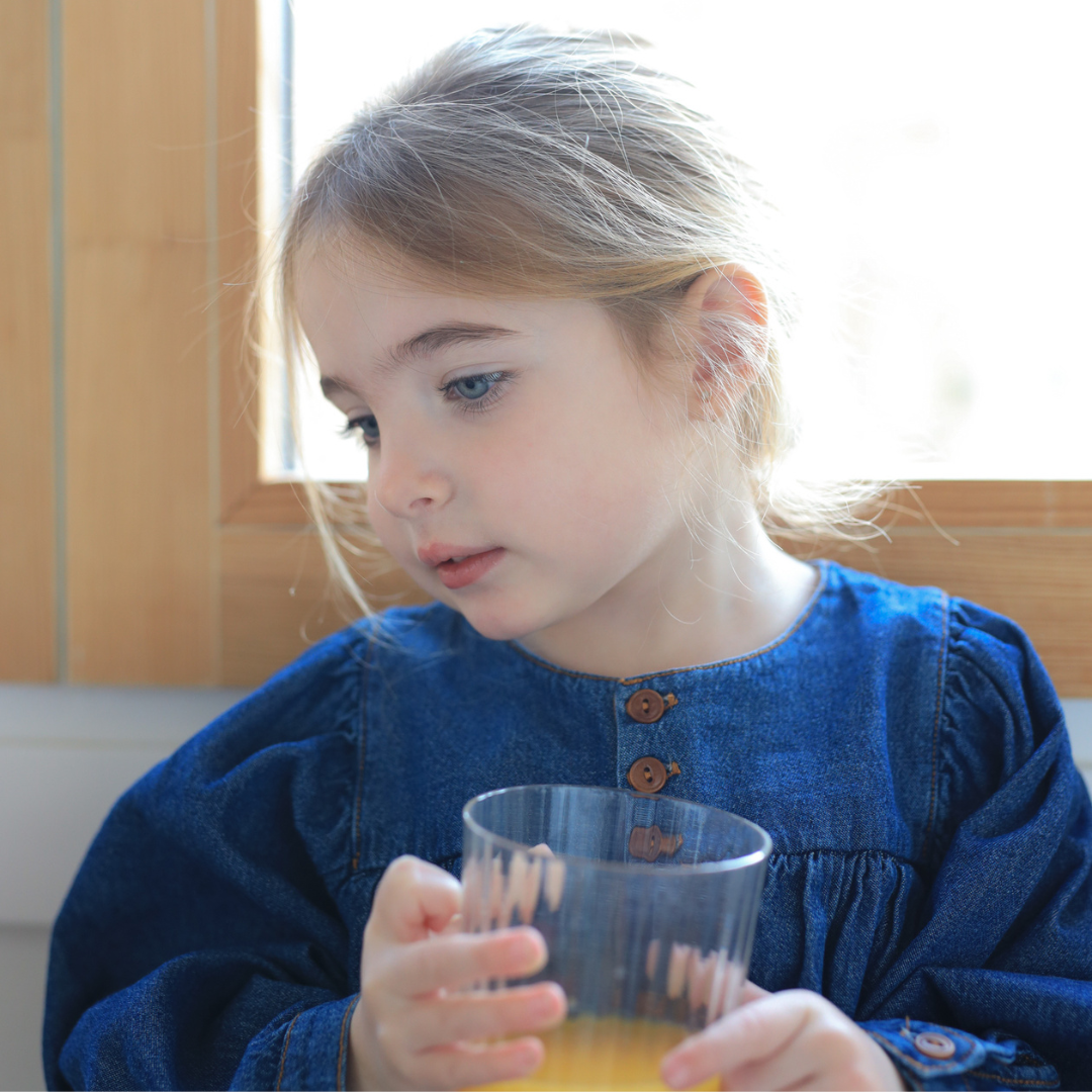 Sold at Bombon shop, designed by 1+ in the family — soft denim baby dress with long sleeves, wooden buttons, and gathered waist. model