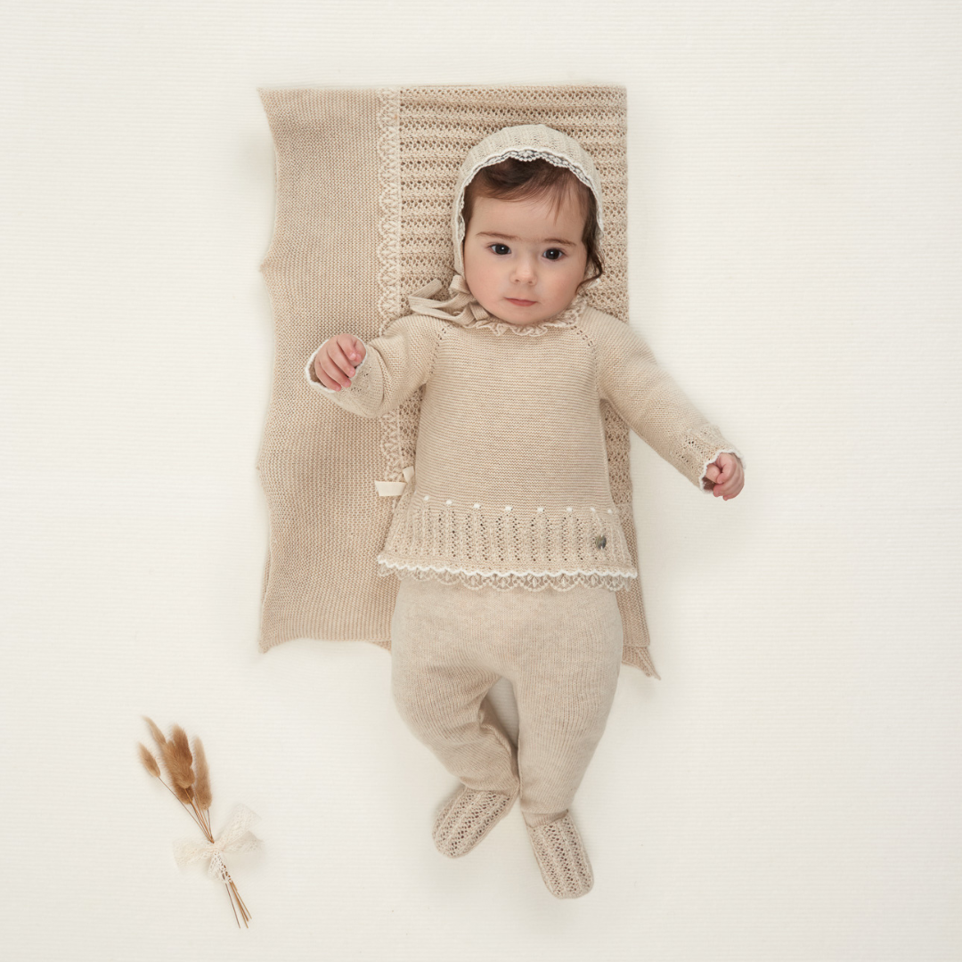 Sold at Bombon shop, designed by Paz Rodriguez. Baby girl beige knit two-piece set with lace trim, pointelle knit, ruffled collar, and footed pants.
