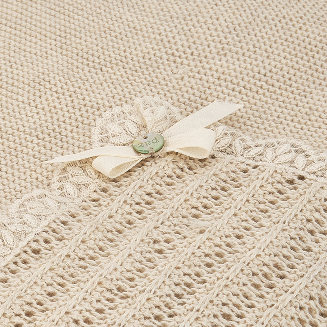 Sold at Bombon shop, designed by Paz Rodriguez: folded beige knit baby blanket with lace trim and pink bow detail.