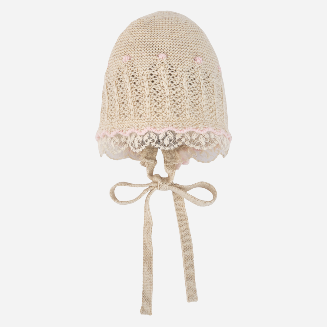 Sold at Bombon shop, designed by Paz Rodriguez: beige knit baby bonnet with lace trim and pink accents.