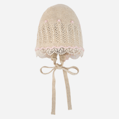 Sold at Bombon shop, designed by Paz Rodriguez: beige knit baby bonnet with lace trim and pink accents.