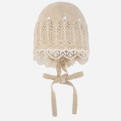 Sold at Bombon shop, designed by Paz Rodriguez, beige knit baby bonnet with lace trim and tie closure.