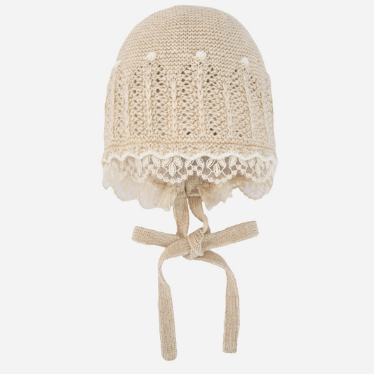Sold at Bombon shop, designed by Paz Rodriguez, beige knit baby bonnet with lace trim and tie closure.