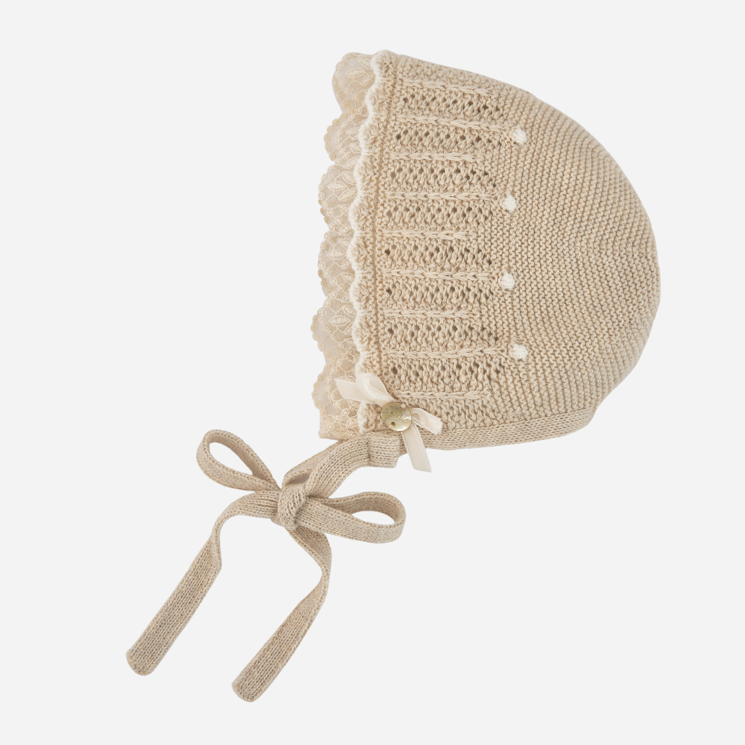 Sold at Bombon shop, designed by Paz Rodriguez, beige knit baby bonnet with lace trim and tie closure.