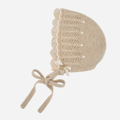 Sold at Bombon shop, designed by Paz Rodriguez, beige knit baby bonnet with lace trim and tie closure.