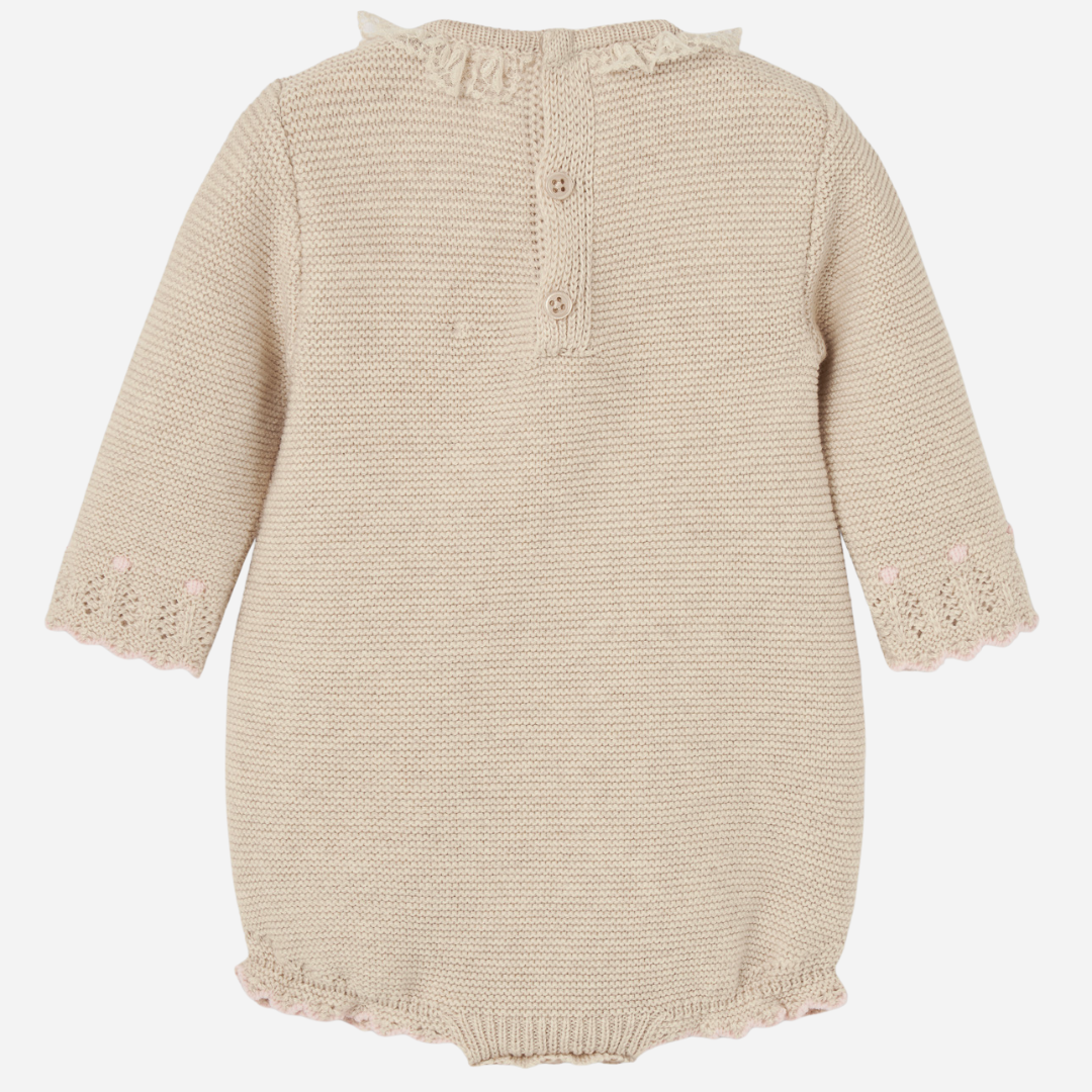 Sold at Bombon shop, designed by Paz Rodriguez, beige knit baby romper with lace detail, scalloped trim, and a pink bow on the waist.