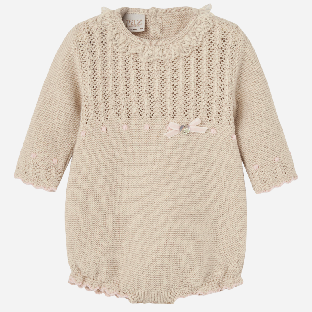 Sold at Bombon shop, designed by Paz Rodriguez, beige knit baby romper with lace detail, scalloped trim, and a pink bow on the waist.