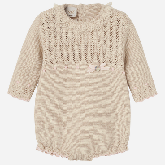 Sold at Bombon shop, designed by Paz Rodriguez, beige knit baby romper with lace detail, scalloped trim, and a pink bow on the waist.