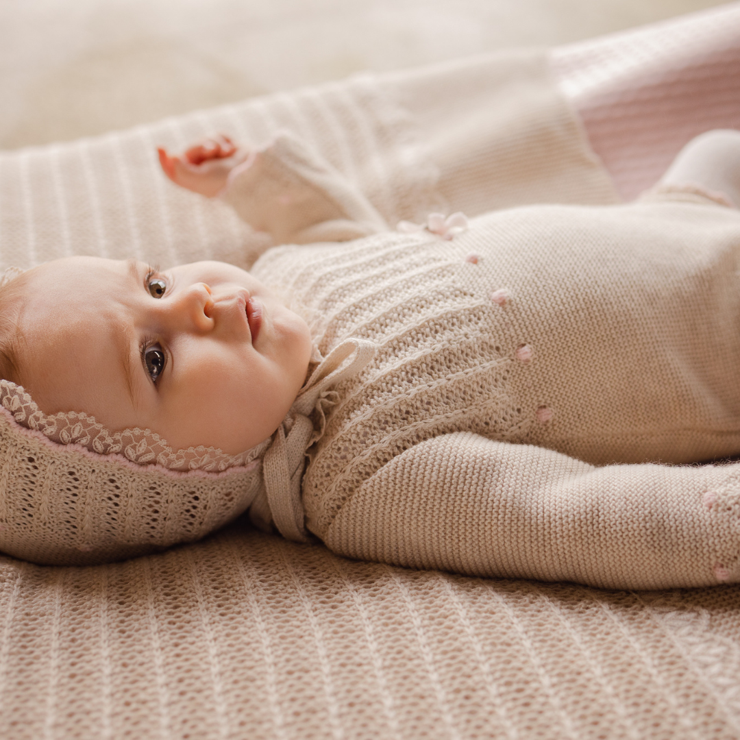 Sold at Bombon shop, designed by Paz Rodriguez, beige knit baby romper with lace detail, scalloped trim, and a pink bow on the waist.