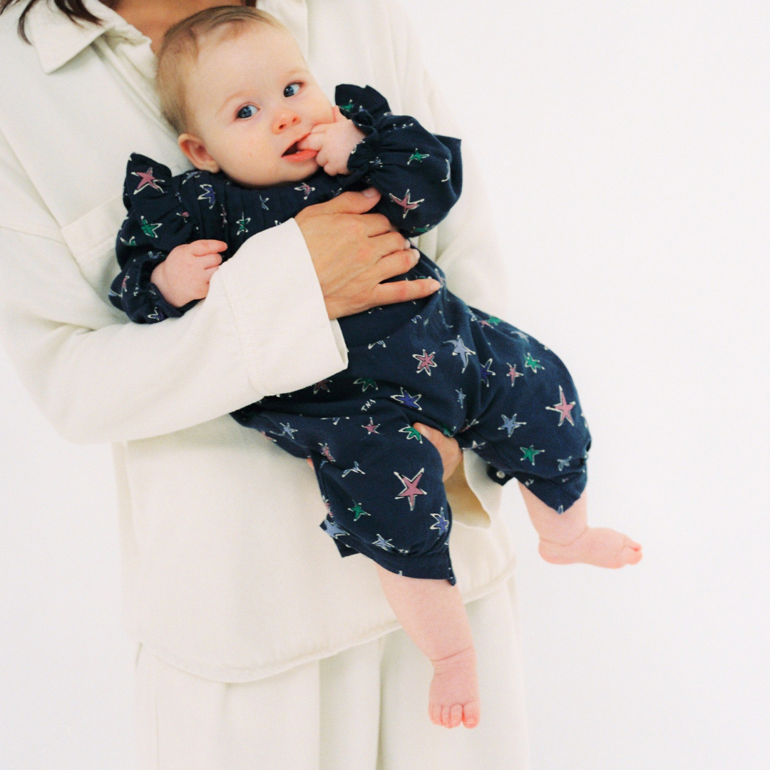 Sold at Bombon shop, this baby girl romper by The New Society features a navy background with a colorful star print, puffed long sleeves, ruffle shoulder details, and gathered cuffs for a whimsical and comfortable all-in-one outfit. model