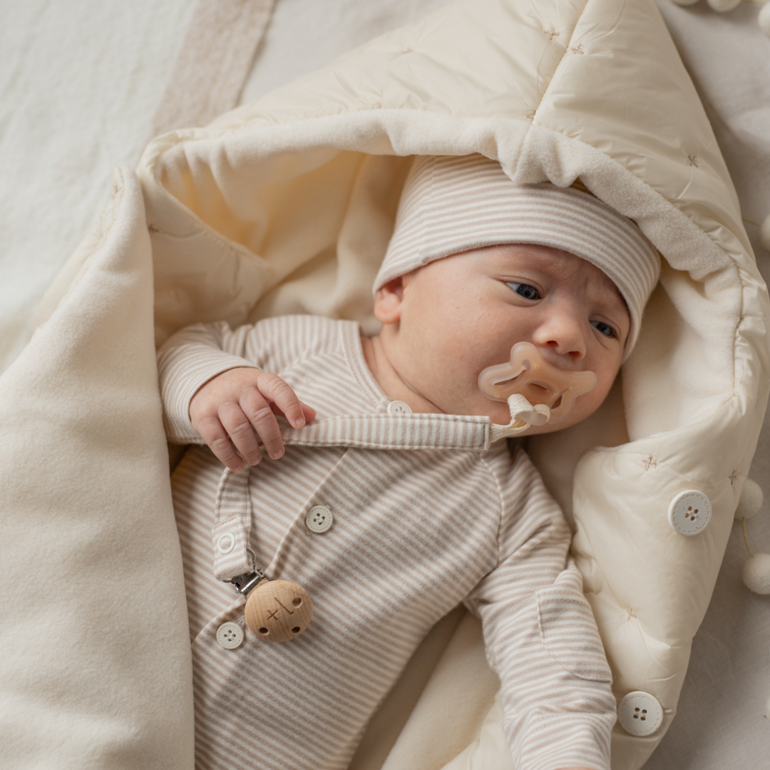Sold at Bombon shop, designed by 1+ in the family — cream-colored quilted baby nest with hood and side button closure, crafted for warmth and comfort. model