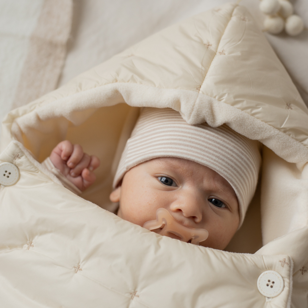Sold at Bombon shop, designed by 1+ in the family — cream-colored quilted baby nest with hood and side button closure, crafted for warmth and comfort. model