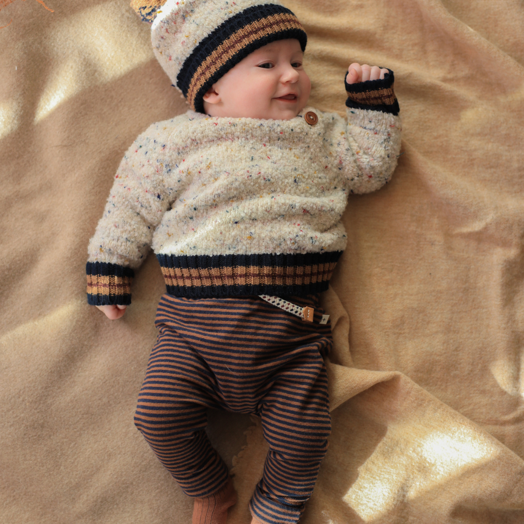 Sold at Bombon shop, designed by 1+ in the family — speckled knit baby sweater with contrast ribbed cuffs and buttoned shoulder, in cream with navy and brown accents. model
