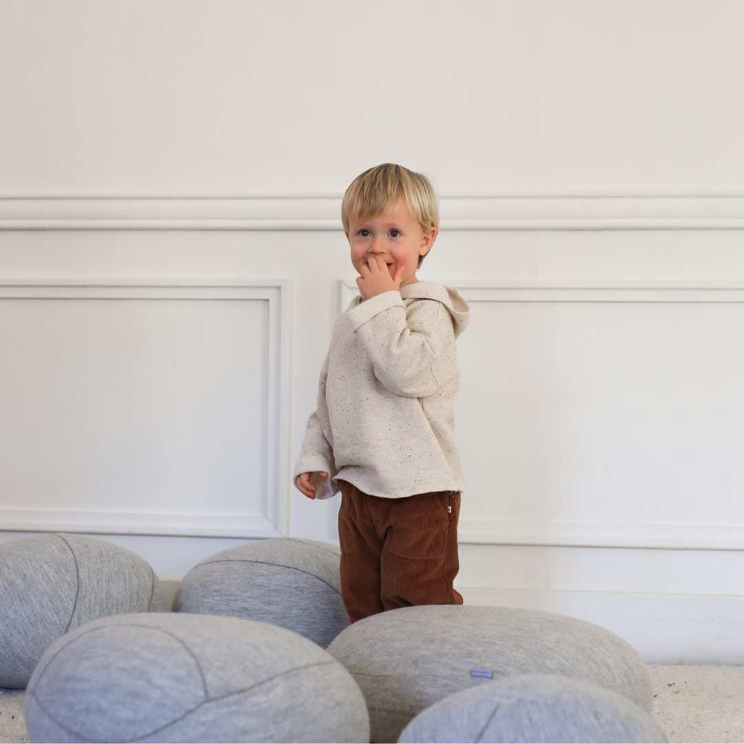 Sold at Bombon shop, designed by 1+ in the family — oatmeal-colored speckled baby hoodie with wooden button and rolled cuffs, photographed flat on white background. model