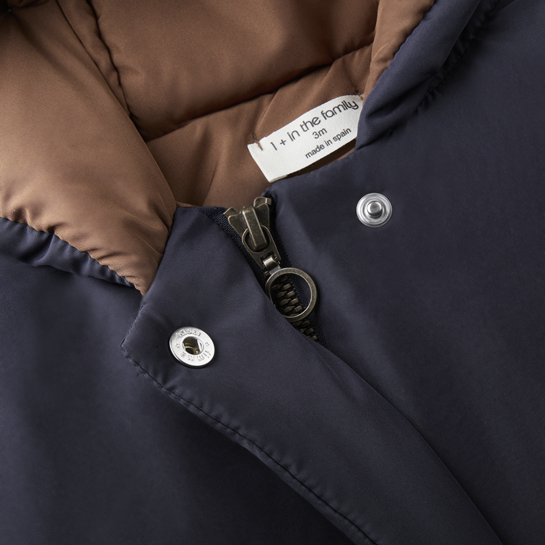 Sold at Bombon shop, designed by 1+ in the family — navy baby puffer jacket with brown lining, hood, front snaps, and patch pockets, laid flat on white background. zoom
