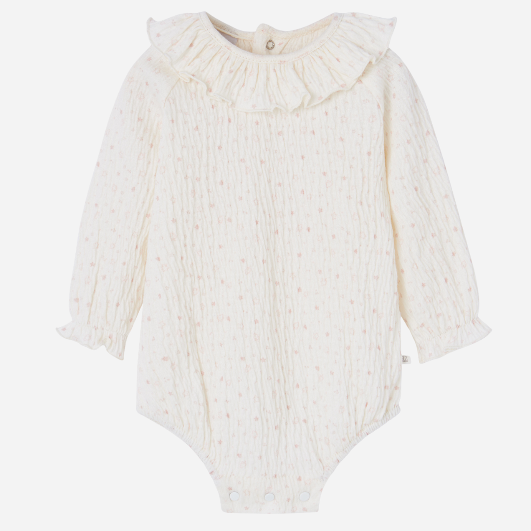 Sold at Bombon shop, designed by Paz Rodriguez. Cream floral muslin baby bodysuit with ruffle collar paired with pink knit overalls featuring front pocket and button straps.