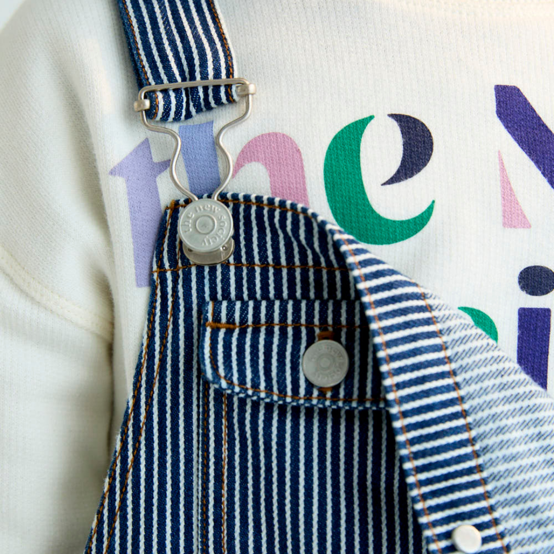 Sold at Bombon Shop, these unisex overalls by The New Society are made from a striped denim with a vintage wash, featuring adjustable shoulder straps, side button closures, and patch pockets for a timeless, relaxed silhouette. detail
