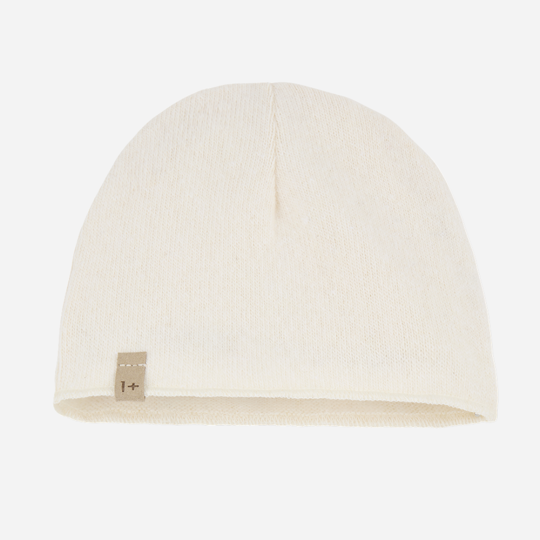 Sold at Bombon shop, designed by 1+ in the family — ecru knit baby beanie with stitched logo tag, laid flat on a white background. front