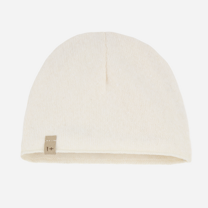Sold at Bombon shop, designed by 1+ in the family — ecru knit baby beanie with stitched logo tag, laid flat on a white background. front