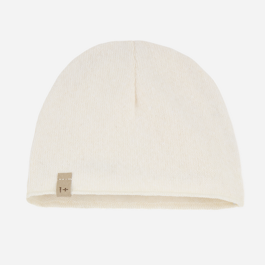 Sold at Bombon shop, designed by 1+ in the family — ecru knit baby beanie with stitched logo tag, laid flat on a white background. front