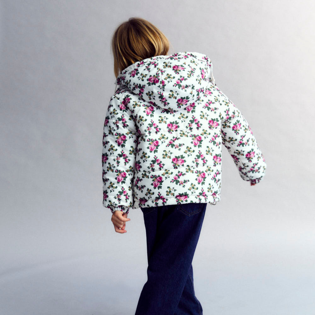 Sold at Bombon shop, this girls’ puffer jacket by The New Society features an all-over rose floral print on a cream base, complete with a hood, front pockets, and snap-button closure—combining warmth, function, and a touch of charm for cooler days. model