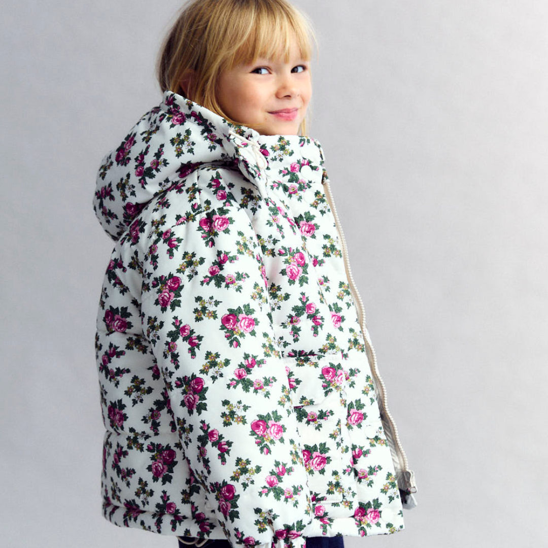 Sold at Bombon shop, this girls’ puffer jacket by The New Society features an all-over rose floral print on a cream base, complete with a hood, front pockets, and snap-button closure—combining warmth, function, and a touch of charm for cooler days. model