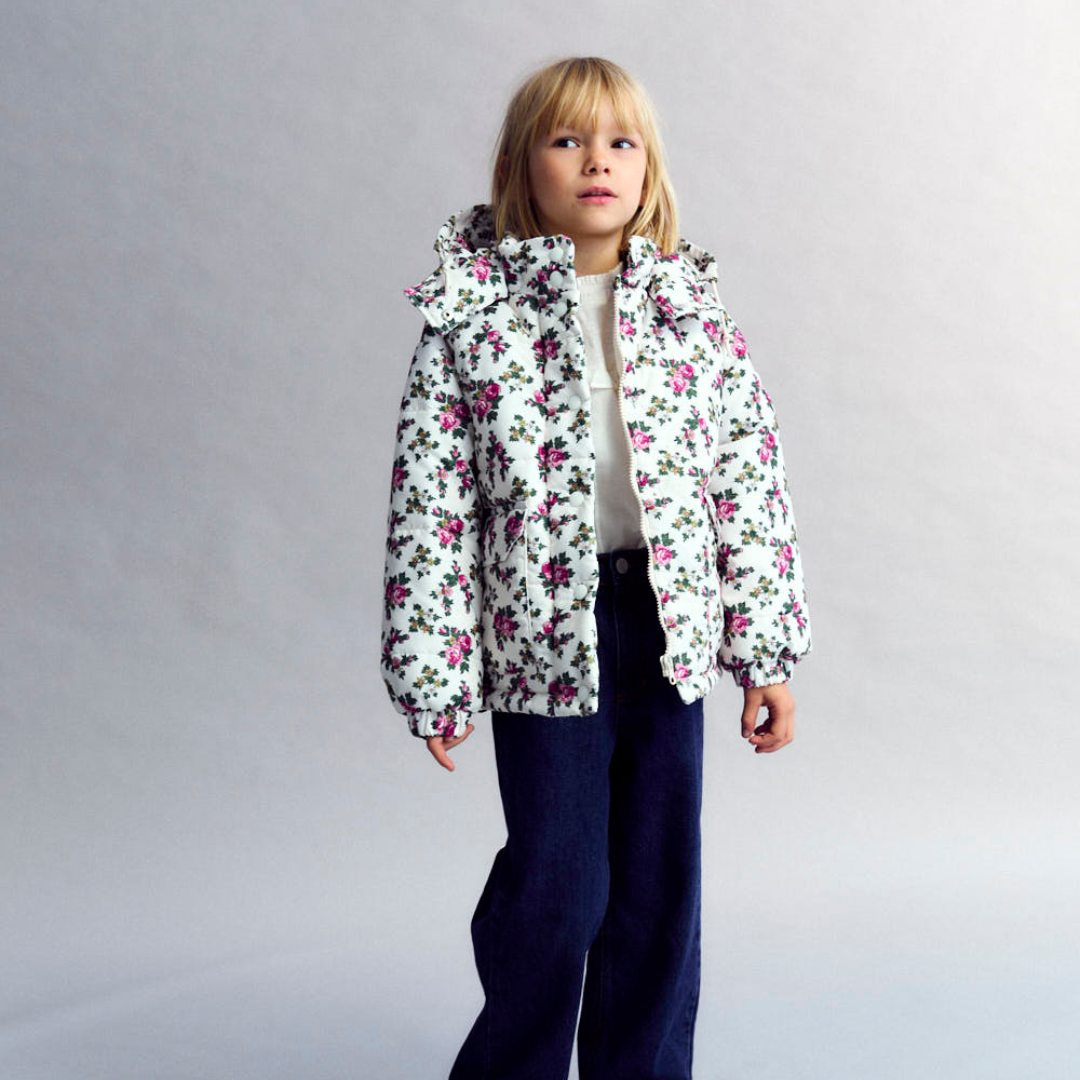 Sold at Bombon shop, this girls’ puffer jacket by The New Society features an all-over rose floral print on a cream base, complete with a hood, front pockets, and snap-button closure—combining warmth, function, and a touch of charm for cooler days. model