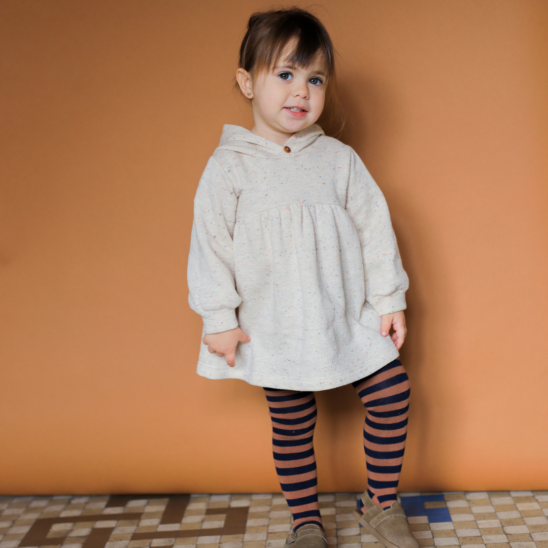 Sold at Bombon shop, designed by 1+ in the family — long-sleeve speckled beige baby dress with gathered waist and hood, shown flat on white background. model