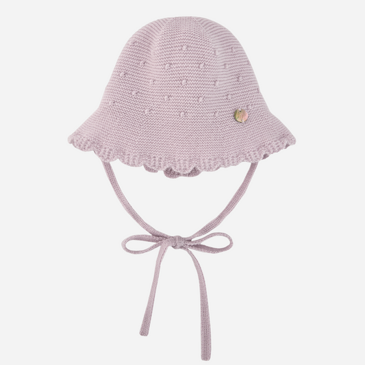 Sold at Bombon shop, designed by Paz Rodriguez, light pink knit baby hat with scalloped brim, bobble texture, and tie closure under the chin.