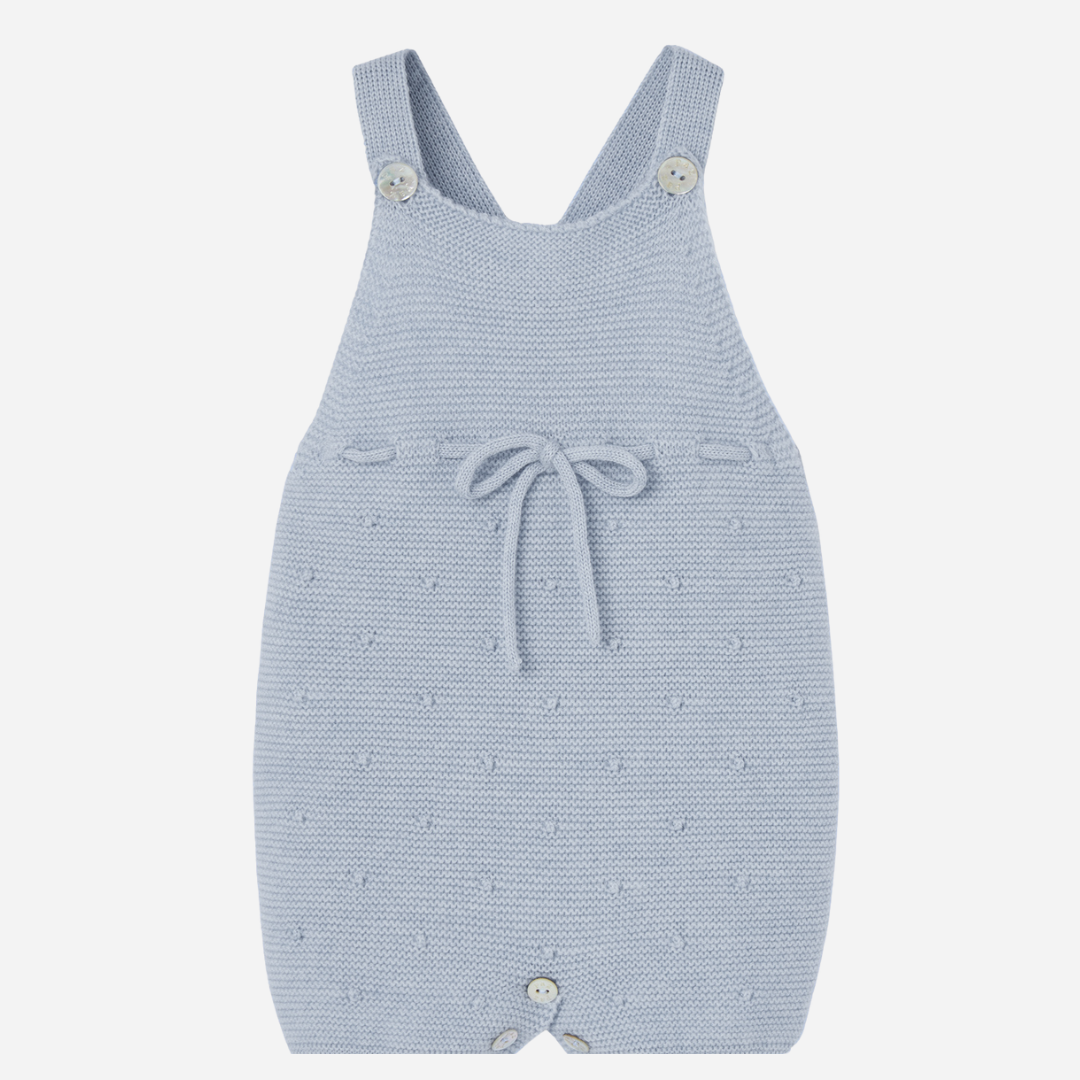 Sold at Bombon shop, designed by Paz Rodriguez. Light blue knitted baby overall with textured dots, waist bow, and button closures.
