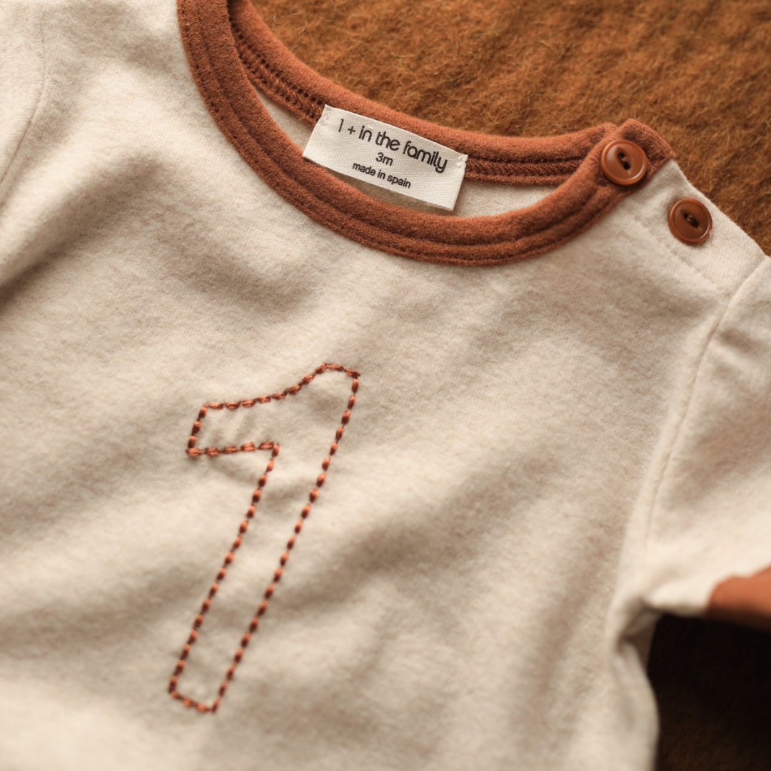 Close-up of a baby outfit with a number '1' embroidered on it, featuring a label on the collar.