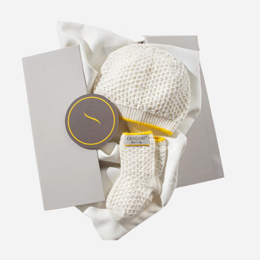Sold at Bombon and designed by Gensami, an ivory newborn layette set including a knit jumpsuit, hat, booties, and blanket with yellow trim
