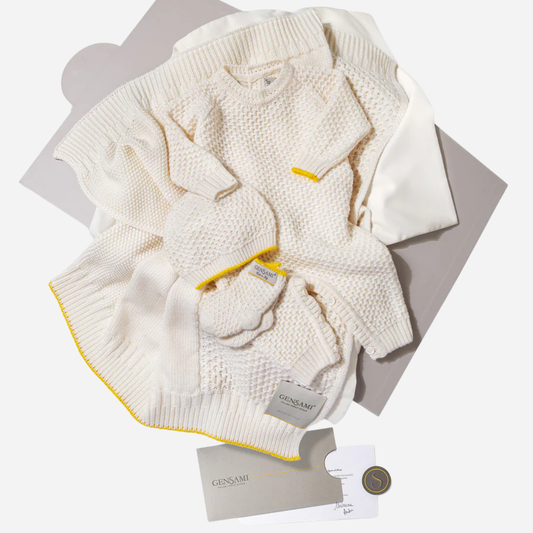 Sold at Bombon and designed by Gensami, an ivory newborn layette set including a knit jumpsuit, hat, booties, and blanket with yellow trim