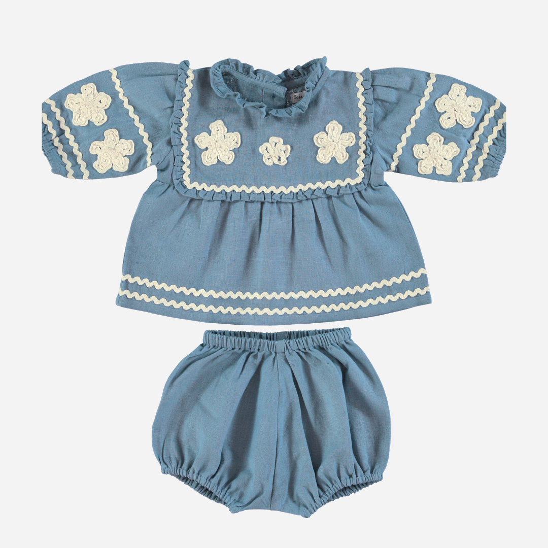 Sold at Bombon Shop, this Bebe Organic baby girl outfit features a vintage-inspired blue top with ivory floral appliqués, ruffled collar, and ric-rac trim, paired with matching bloomers — a timeless and elegant set perfect for special occasions or everyday charm.