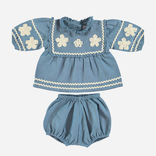 Sold at Bombon Shop, this Bebe Organic baby girl outfit features a vintage-inspired blue top with ivory floral appliqués, ruffled collar, and ric-rac trim, paired with matching bloomers — a timeless and elegant set perfect for special occasions or everyday charm.